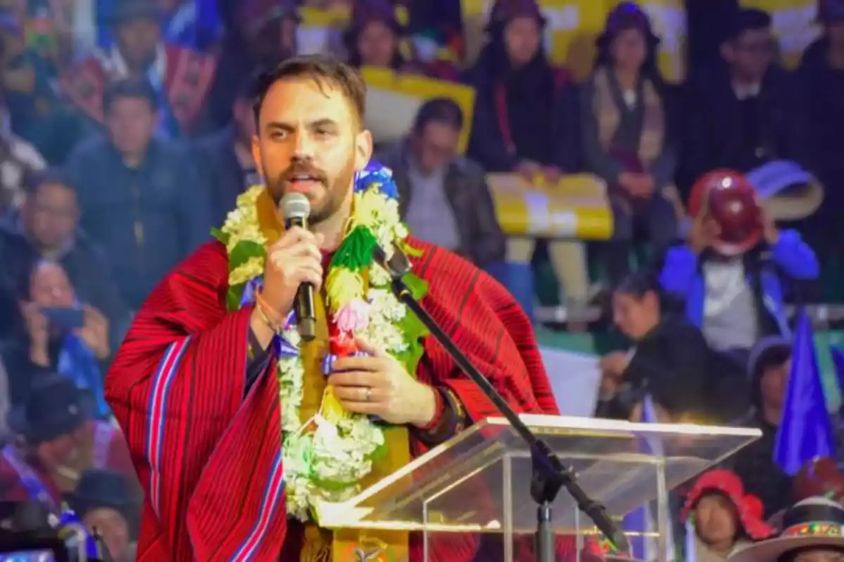 Hombre con poncho rojo y guirnalda de flores hablando en un micrófono detrás de un atril transparente frente a una multitud