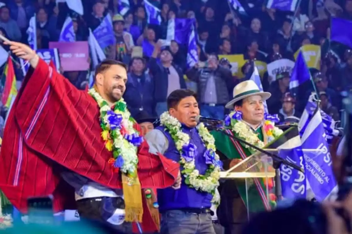 Tres hombres con guirnaldas de flores y ponchos coloridos en un escenario durante un evento político con banderas y público al fondo