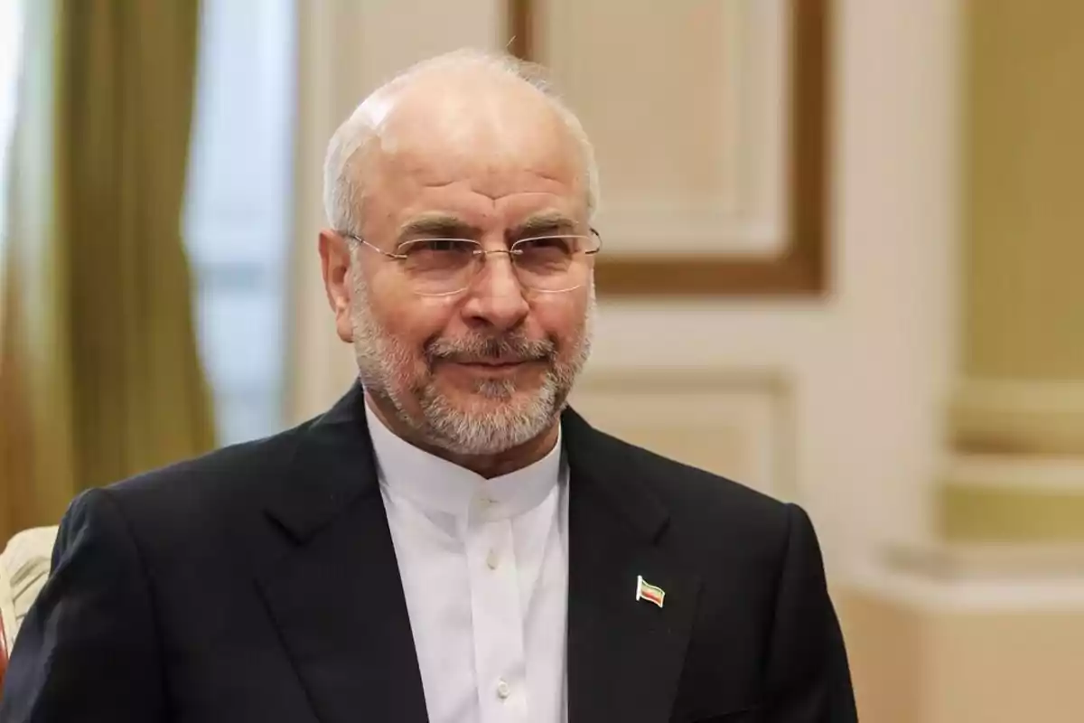 Middle-aged man with a beard and glasses, dressed in a dark suit and white jersey, wearing a pin with the Iranian flag on his lapel, in an elegant indoor setting. Middle-aged man with a beard and glasses, dressed in a dark suit and white jersey, wearing a pin with the Iranian flag on his lapel, in an elegant indoor setting.