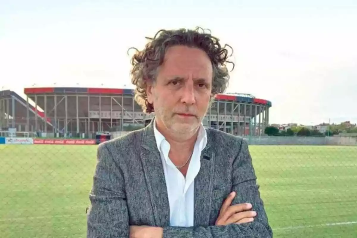 Curly-haired, gray-haired man wearing a gray jacket and white jersey standing on a soccer field with a stadium in the background Curly-haired, gray-haired man wearing a gray jacket and white jersey standing on a soccer field with a stadium in the background