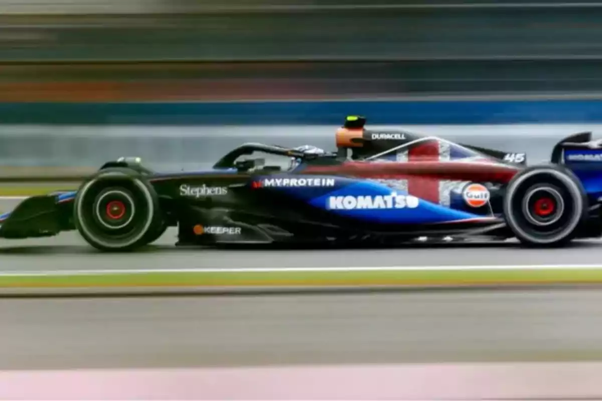 Un monoplaza de Fórmula 1 con decoración azul, negra y detalles de la bandera británica circula a gran velocidad en una pista de carreras.