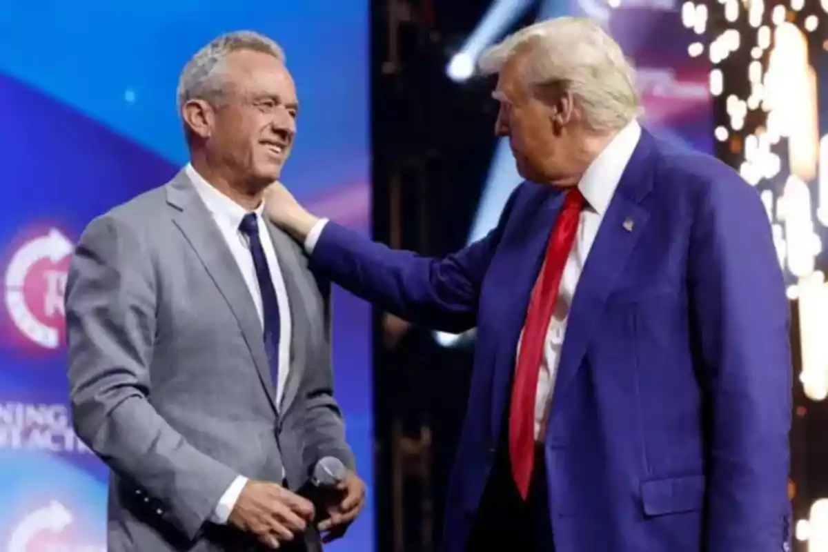 Two men in suits are talking on a lit stage while one of them holds a microphone and the other puts his hand on his shoulder.