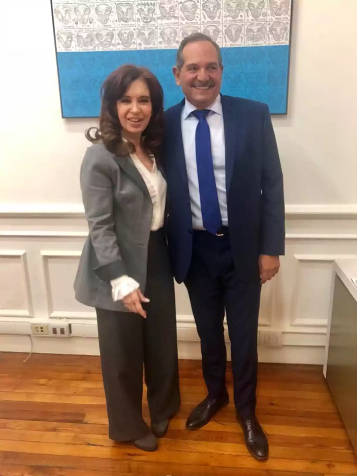 Dos personas posan sonrientes en una oficina elegante con piso de madera y una pintura azul y blanca en la pared. Dos personas posan sonrientes en una oficina elegante con piso de madera y una pintura azul y blanca en la pared.