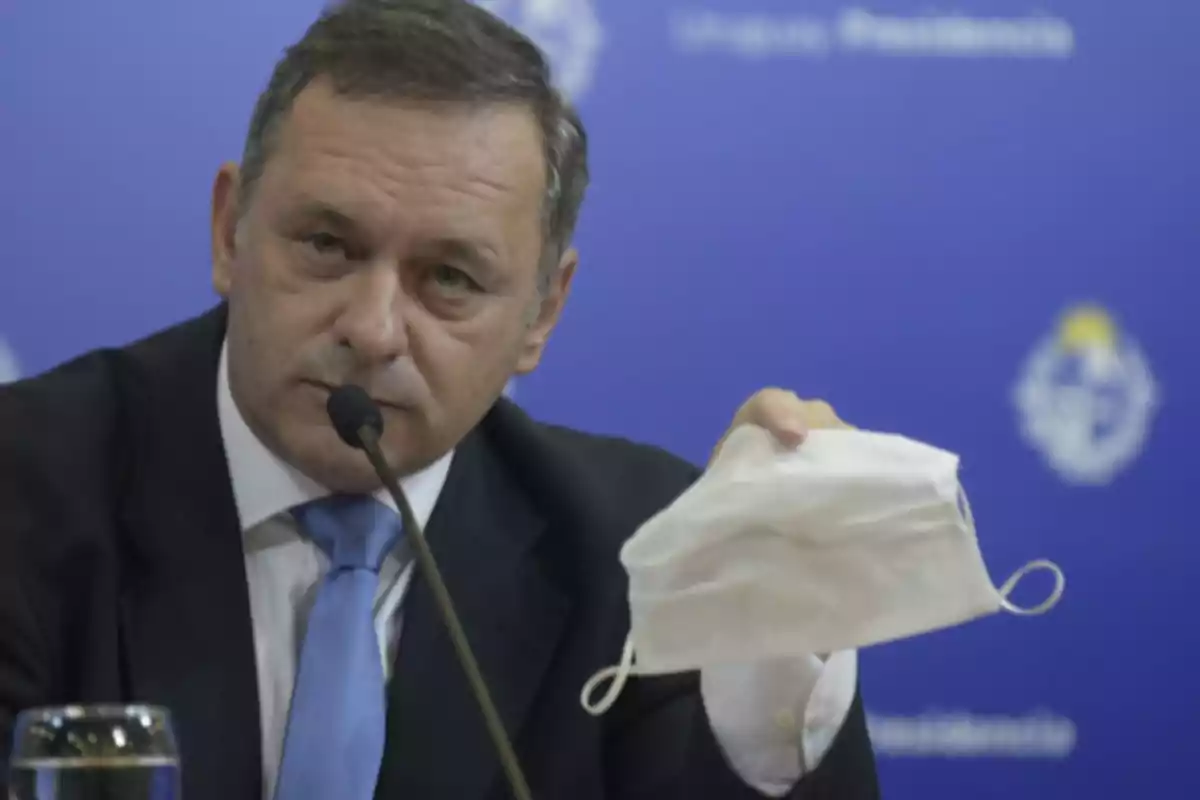 Hombre de traje y corbata azul sosteniendo un tapabocas blanco mientras habla frente a un micrófono con fondo azul institucional Hombre de traje y corbata azul sosteniendo un tapabocas blanco mientras habla frente a un micrófono con fondo azul institucional