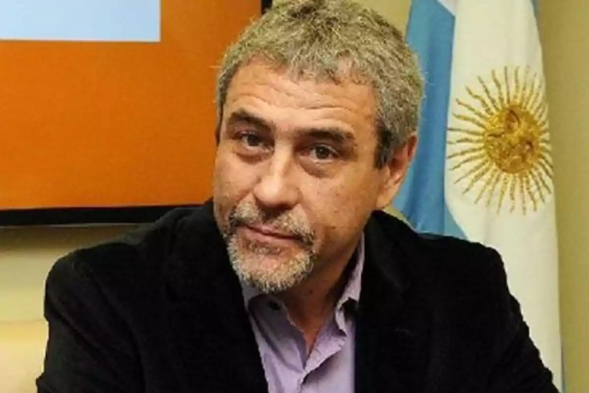 Gray-haired man with a short beard wearing a dark jacket and a lilac jersey sitting in front of an Argentine flag Gray-haired man with a short beard wearing a dark jacket and a lilac jersey sitting in front of an Argentine flag