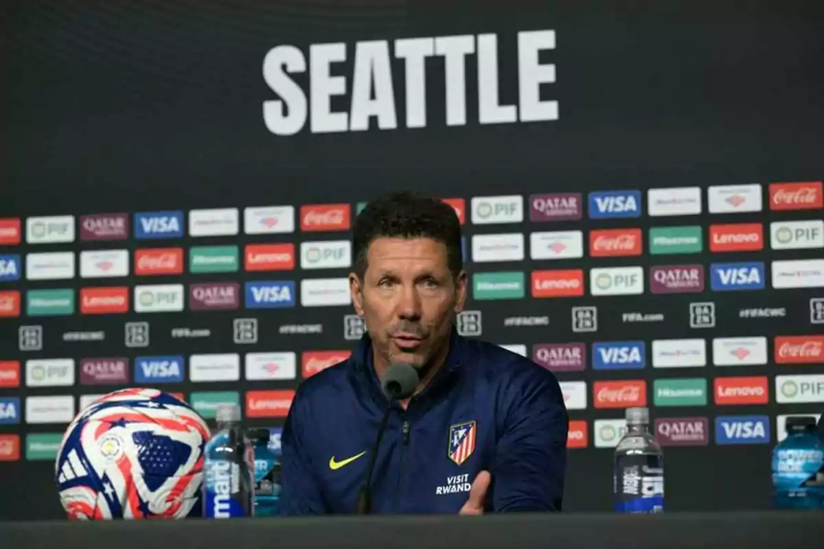 Atlético de Madrid coach speaking at a press conference in Seattle with a ball and water bottles on the table and a backdrop with sponsor logos. Atlético de Madrid coach speaking at a press conference in Seattle with a ball and water bottles on the table and a backdrop with sponsor logos.