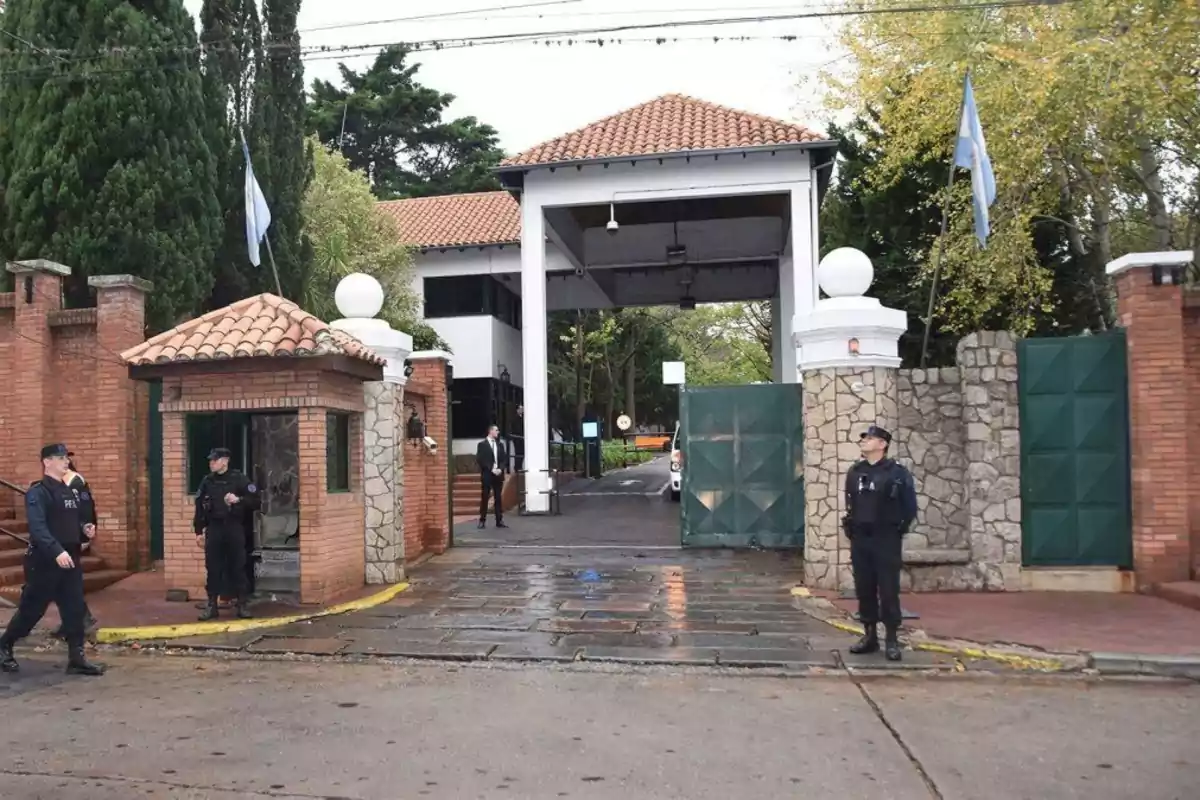 Main entrance of a residence guarded by several police officers, with Argentine flags waving and an open green gate