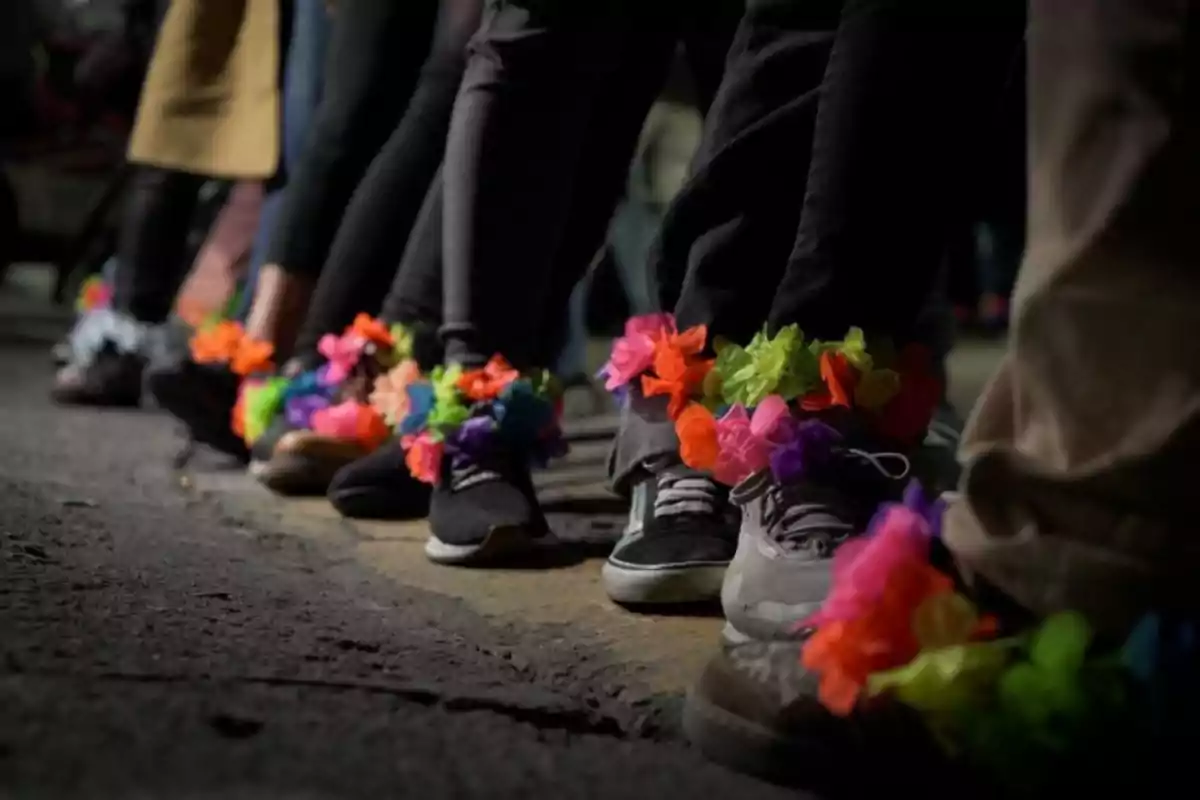 Personas alineadas usando tenis decorados con flores de papel de colores en los tobillos sobre una calle