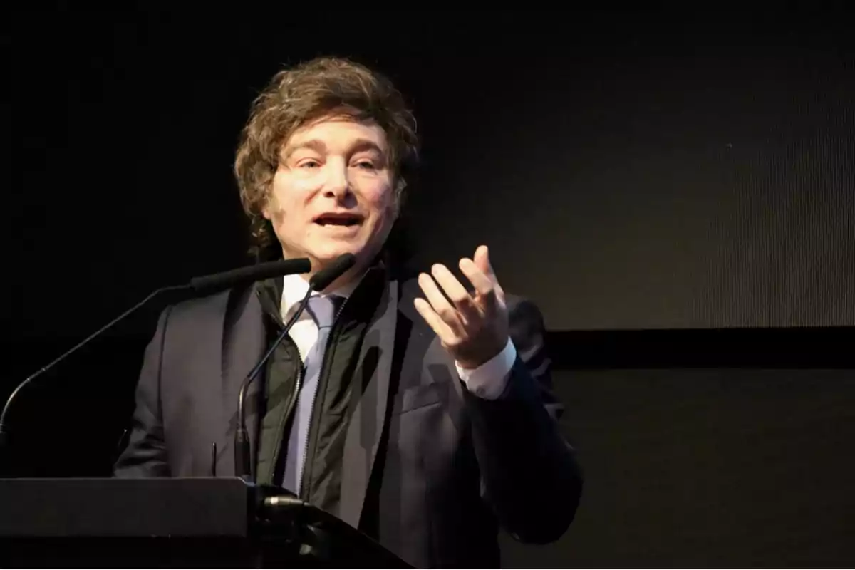 Brown-haired man in a dark suit speaking at a podium with a microphone and gesturing with his hand
