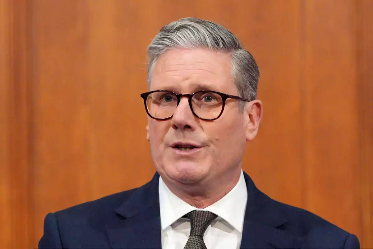 Gray-haired man with glasses in a dark suit and tie speaking in front of a wooden background Gray-haired man with glasses in a dark suit and tie speaking in front of a wooden background