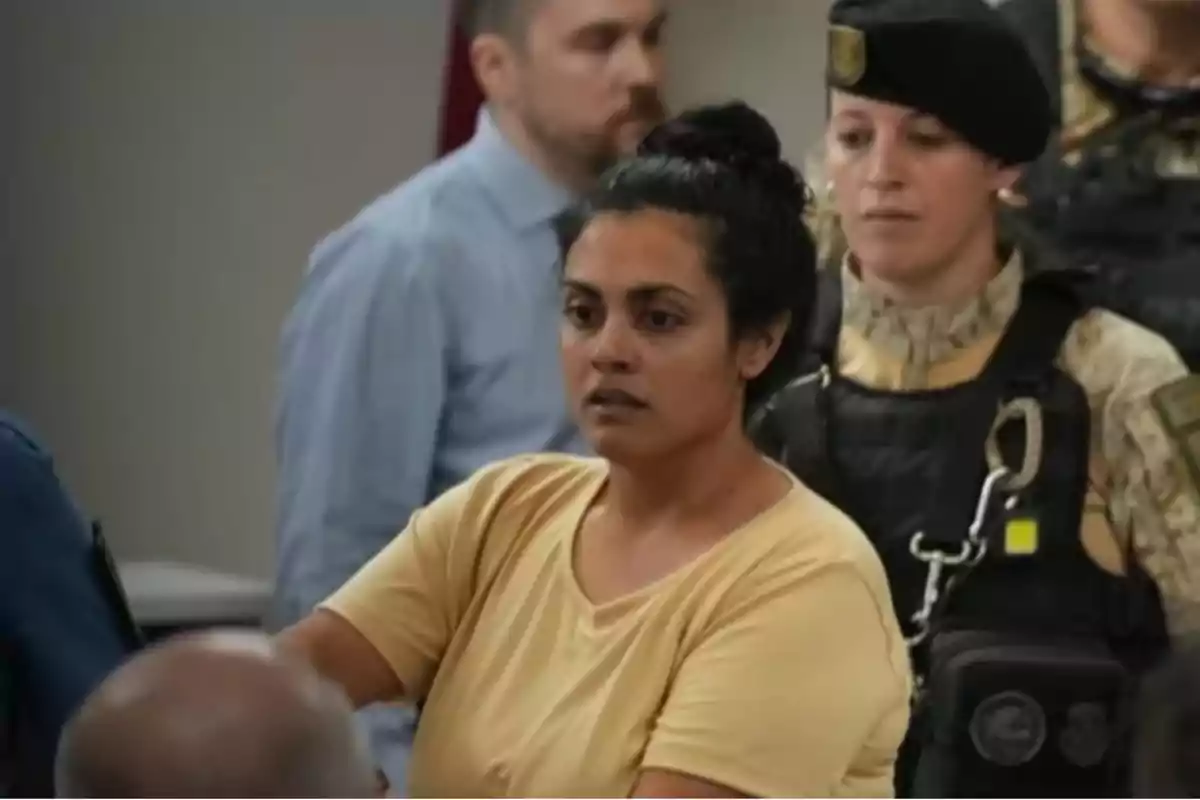 Mujer de cabello oscuro recogido y camiseta amarilla está escoltada por una oficial con uniforme táctico en un entorno interior con otras personas al fondo
