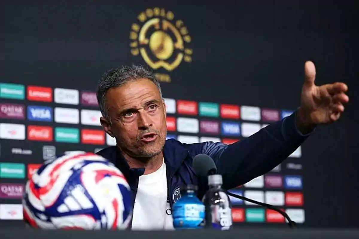 Soccer coach speaking at a press conference with a ball and water bottles on the table and a backdrop with sponsor logos.
