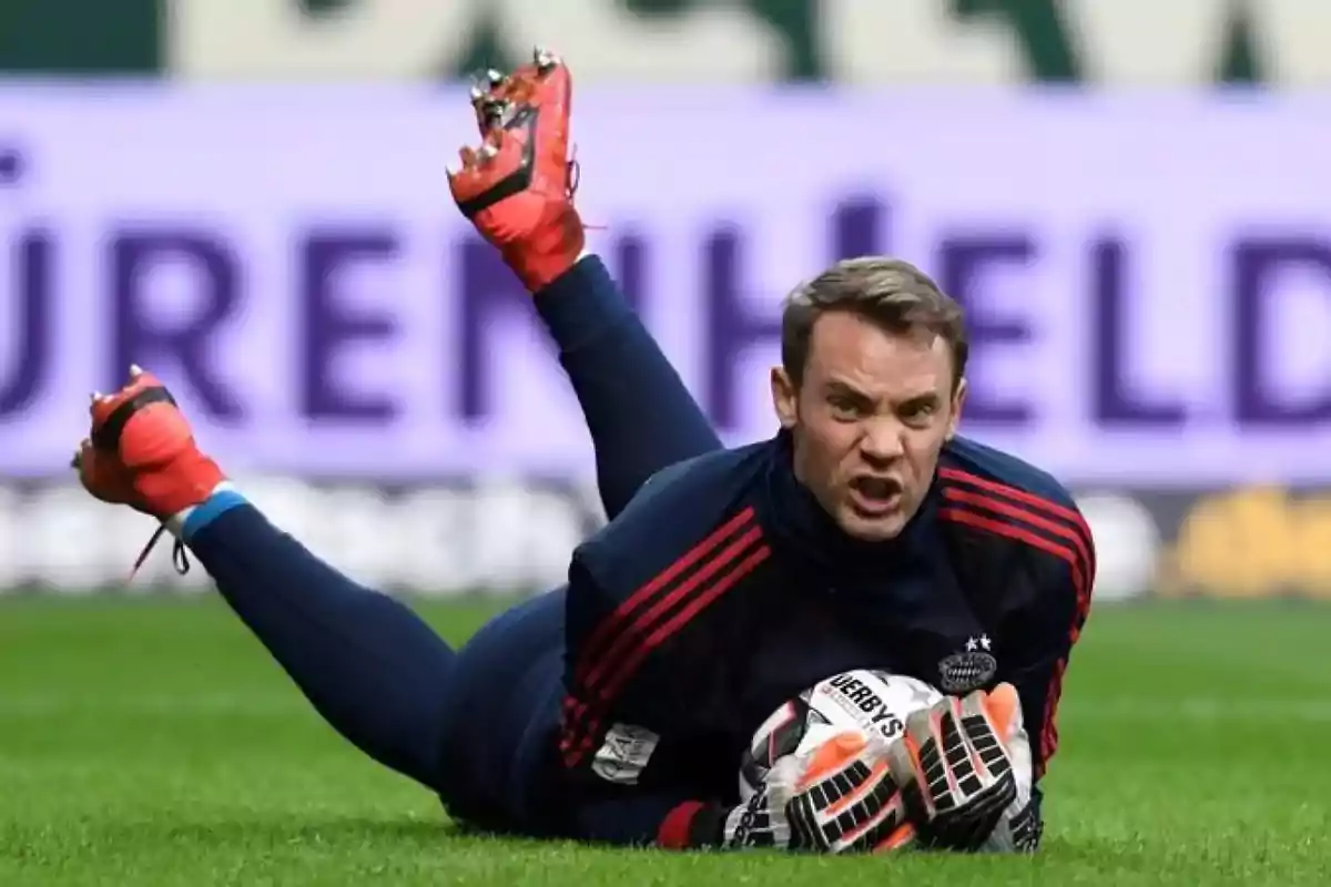Portero de fútbol con uniforme oscuro y guantes rojos detiene el balón en el césped mientras hace una expresión de esfuerzo Portero de fútbol con uniforme oscuro y guantes rojos detiene el balón en el césped mientras hace una expresión de esfuerzo
