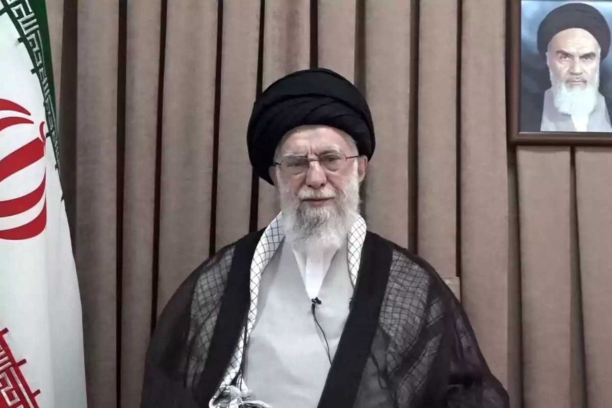 An elderly man with a white beard and a black turban is sitting in front of a beige curtain, next to an Iranian flag and a portrait on the wall. An elderly man with a white beard and a black turban is sitting in front of a beige curtain, next to an Iranian flag and a portrait on the wall.