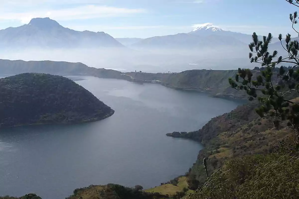 Vista panorámica de una laguna rodeada de montañas con vegetación y un volcán nevado al fondo bajo un cielo despejado Vista panorámica de una laguna rodeada de montañas con vegetación y un volcán nevado al fondo bajo un cielo despejado