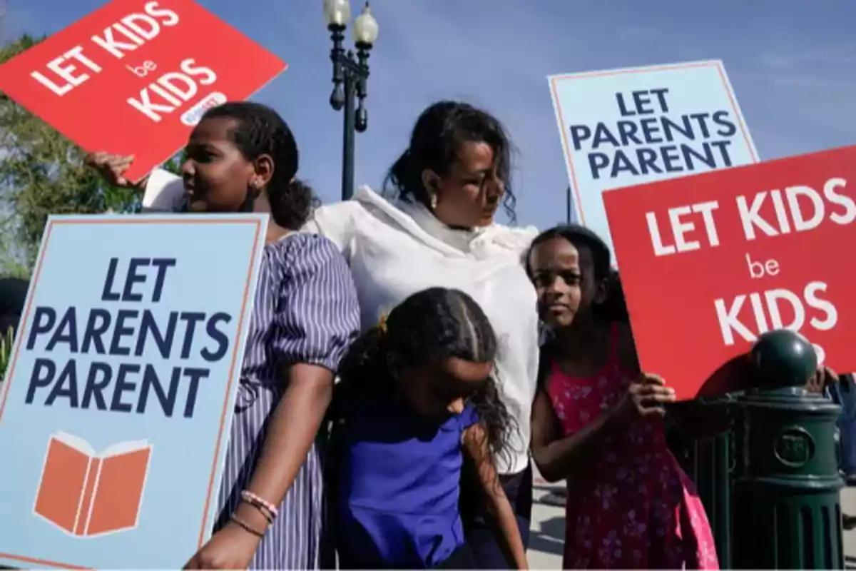 Una mujer y tres niñas sostienen carteles que dicen Let Parents Parent y Let Kids be Kids durante una manifestación al aire libre en un día soleado