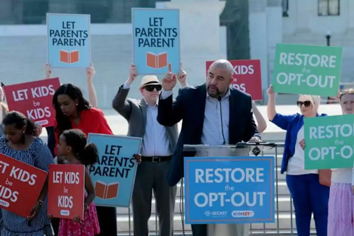 Un grupo de personas sostiene carteles con mensajes como Let Parents Parent, Let Kids be Kids y Restore the Opt-Out mientras un hombre habla en un podio durante una manifestación al aire libre.