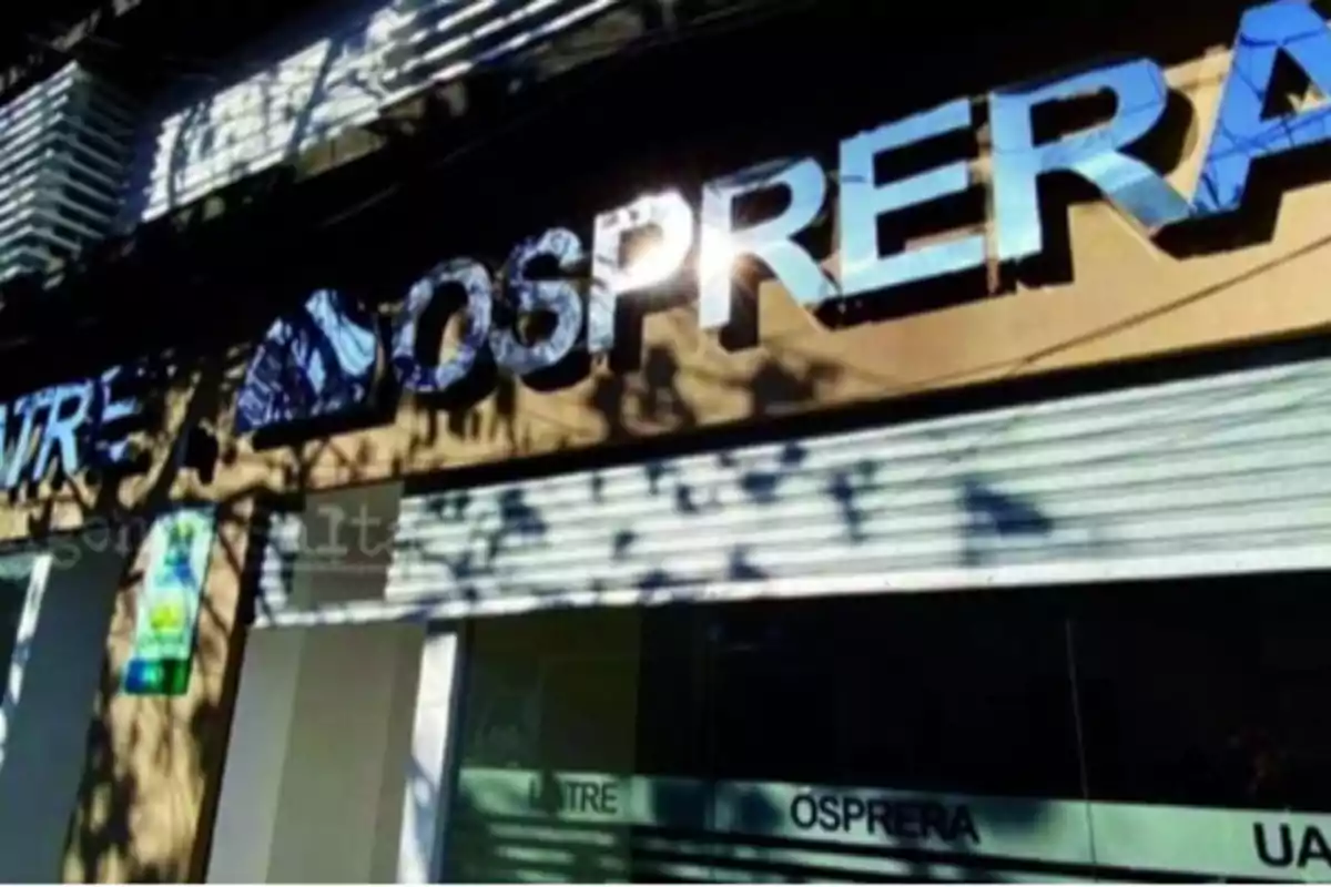 Fachada de un edificio con el cartel de OSPRERA reflejando la luz del sol y sombras de árboles sobre la pared