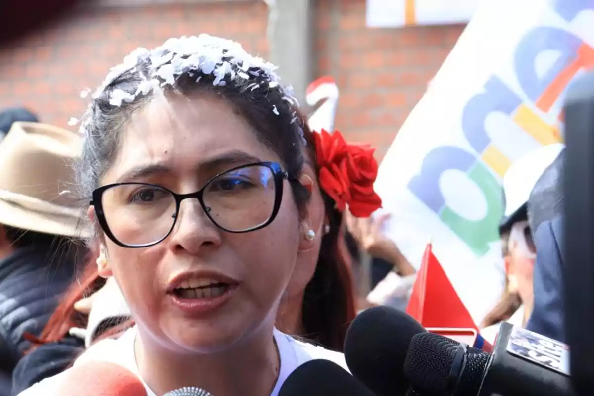 Mujer con gafas y una flor roja en el cabello hablando frente a varios micrófonos en un evento al aire libre Mujer con gafas y una flor roja en el cabello hablando frente a varios micrófonos en un evento al aire libre