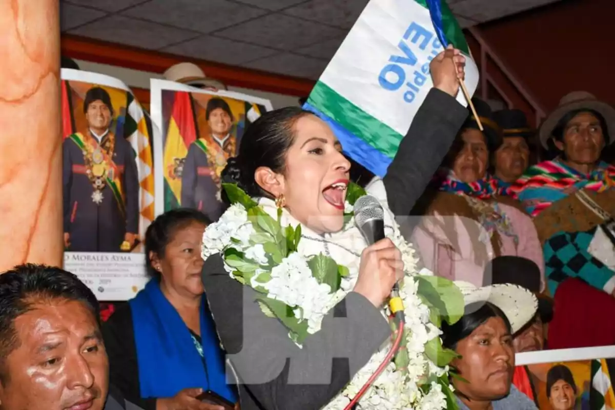 Mujer con guirnalda de flores hablando por micrófono y levantando una bandera en un evento político rodeada de personas y carteles de Evo Morales Mujer con guirnalda de flores hablando por micrófono y levantando una bandera en un evento político rodeada de personas y carteles de Evo Morales
