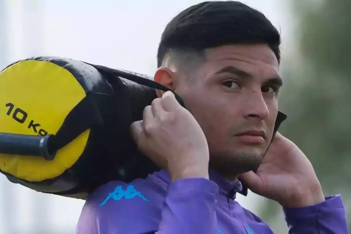 Hombre con cabello corto y barba ligera cargando un saco de entrenamiento amarillo de 10 kilos sobre los hombros mientras usa una sudadera morada