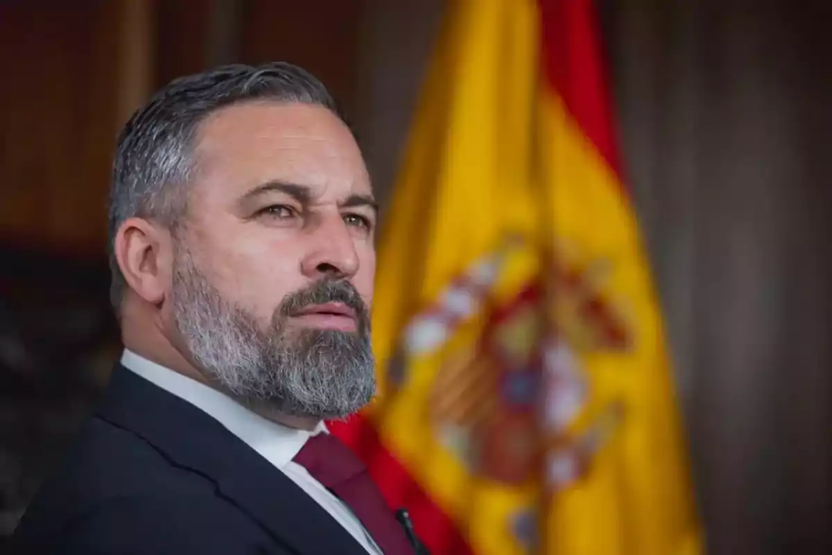 Hombre de barba canosa con traje y corbata roja frente a una bandera de España desenfocada