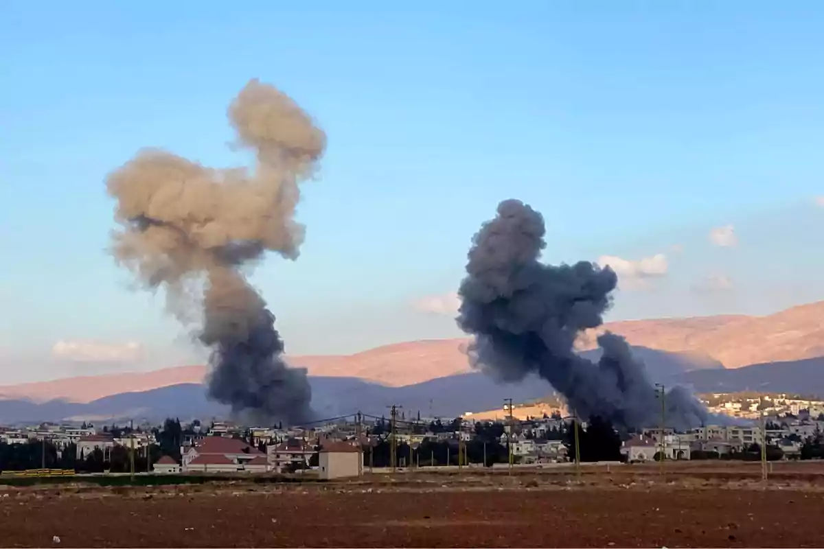 Clouds of gray and black smoke rise over a city after explosions in an urban area surrounded by mountains under a clear sky.