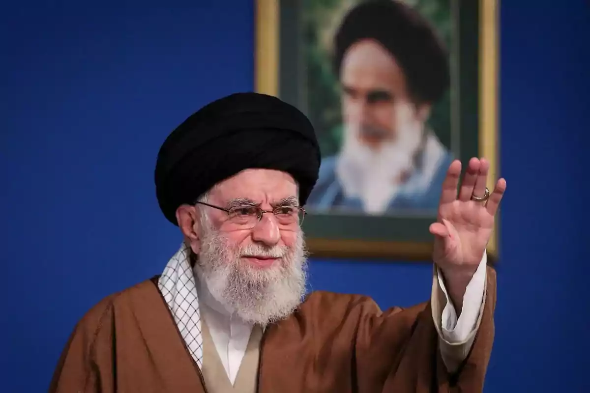 Older man with a beard and glasses dressed in a brown robe and black turban raising his hand in front of a blue background and a blurred portrait