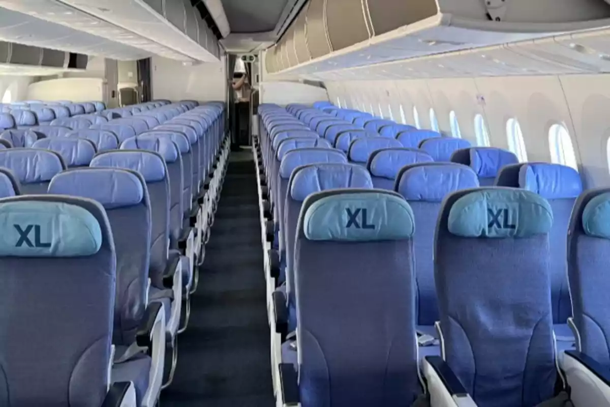Interior de un avión con filas de asientos vacíos tapizados en azul y cabeceras que tienen la inscripción XL Interior de un avión con filas de asientos vacíos tapizados en azul y cabeceras que tienen la inscripción XL