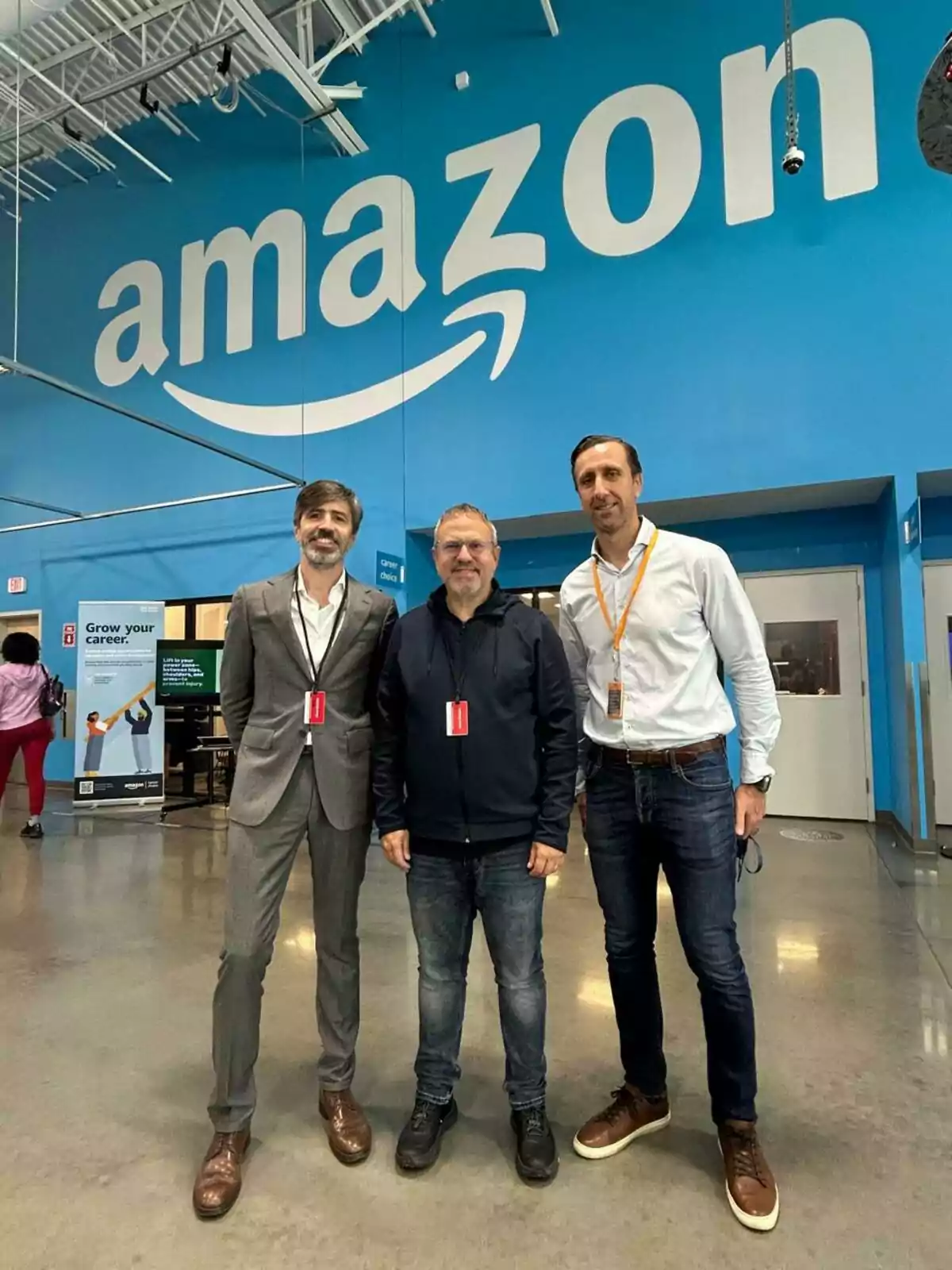 Tres hombres posan juntos en el interior de una instalación de Amazon con una gran pared azul de fondo que muestra el logotipo de la empresa. Tres hombres posan juntos en el interior de una instalación de Amazon con una gran pared azul de fondo que muestra el logotipo de la empresa.