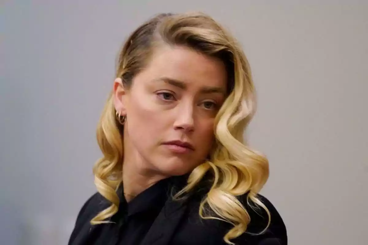 Mujer de cabello rubio ondulado y expresión seria viste ropa oscura y está en un entorno interior con fondo neutro Mujer de cabello rubio ondulado y expresión seria viste ropa oscura y está en un entorno interior con fondo neutro