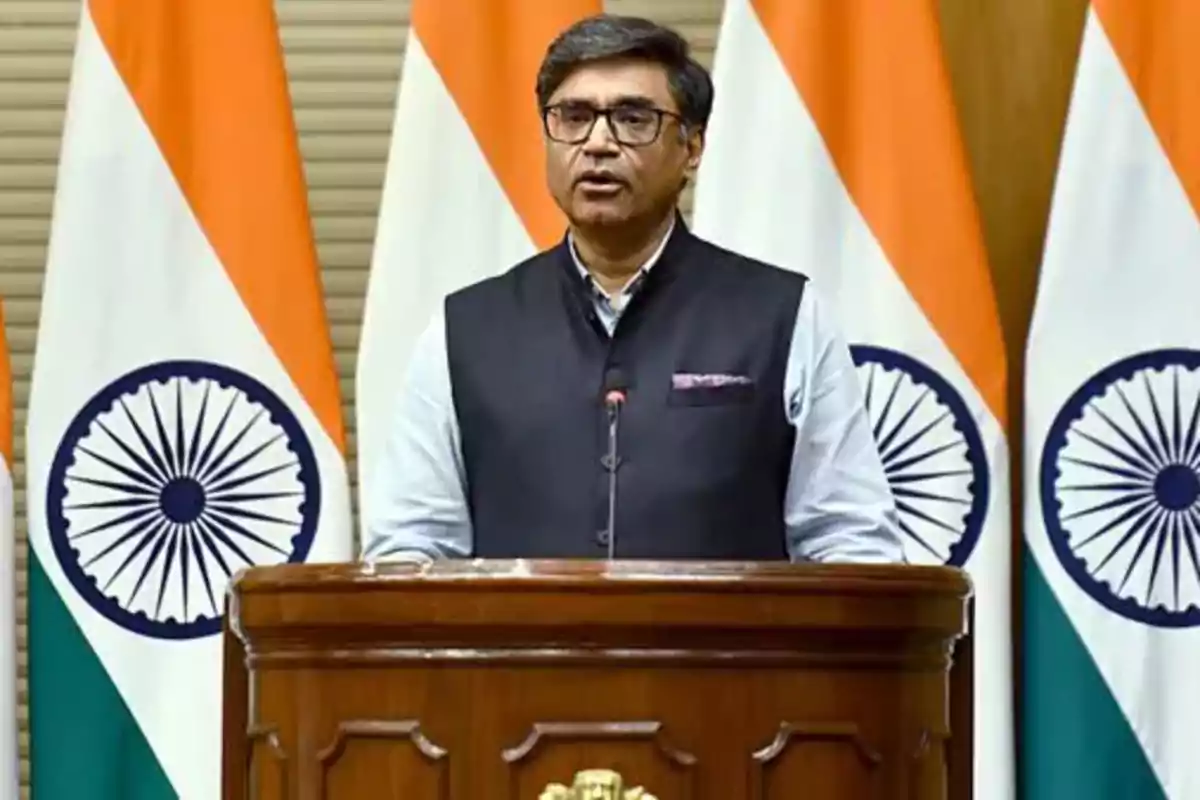 A man wearing glasses and a dark vest speaks at a podium in front of several Indian flags. A man wearing glasses and a dark vest speaks at a podium in front of several Indian flags.