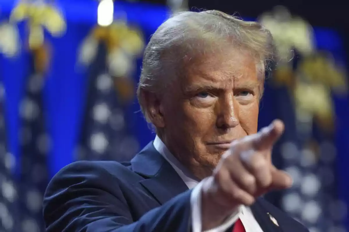 Blond-haired man in a blue suit pointing forward with a serious expression and a background of blurred flags Blond-haired man in a blue suit pointing forward with a serious expression and a background of blurred flags