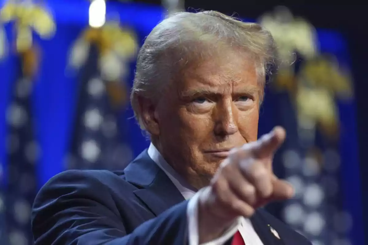 Blond-haired man in a blue suit pointing forward with a serious expression and a background of blurred flags Blond-haired man in a blue suit pointing forward with a serious expression and a background of blurred flags