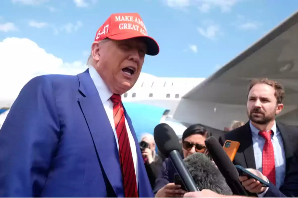 Hombre con gorra roja y traje azul hablando frente a varios micrófonos mientras es entrevistado por periodistas cerca de un avión Hombre con gorra roja y traje azul hablando frente a varios micrófonos mientras es entrevistado por periodistas cerca de un avión