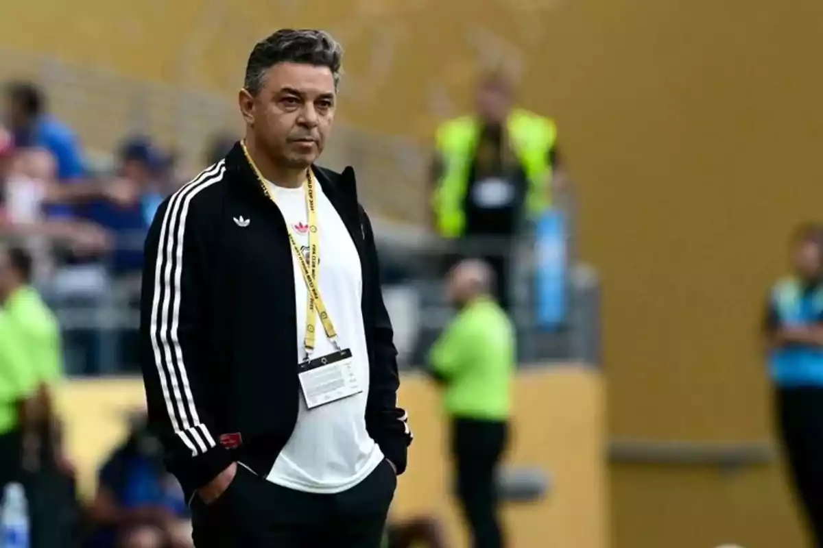 Hombre con chaqueta deportiva negra y camiseta blanca de pie en un estadio con personas desenfocadas al fondo