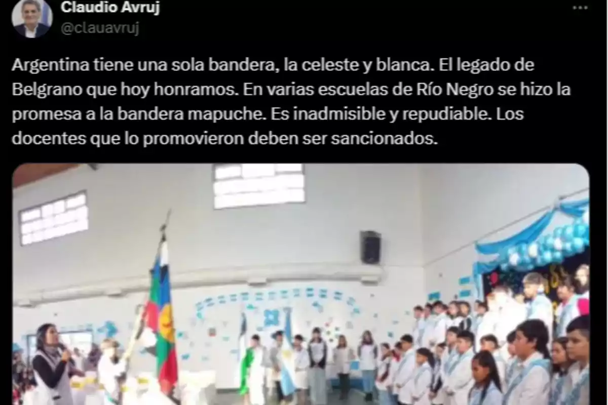Acto escolar en un gimnasio decorado con los colores celeste y blanco, donde un grupo de niños y docentes vestidos con guardapolvos blancos participan de una ceremonia junto a la bandera argentina y la bandera mapuche. Acto escolar en un gimnasio decorado con los colores celeste y blanco, donde un grupo de niños y docentes vestidos con guardapolvos blancos participan de una ceremonia junto a la bandera argentina y la bandera mapuche.