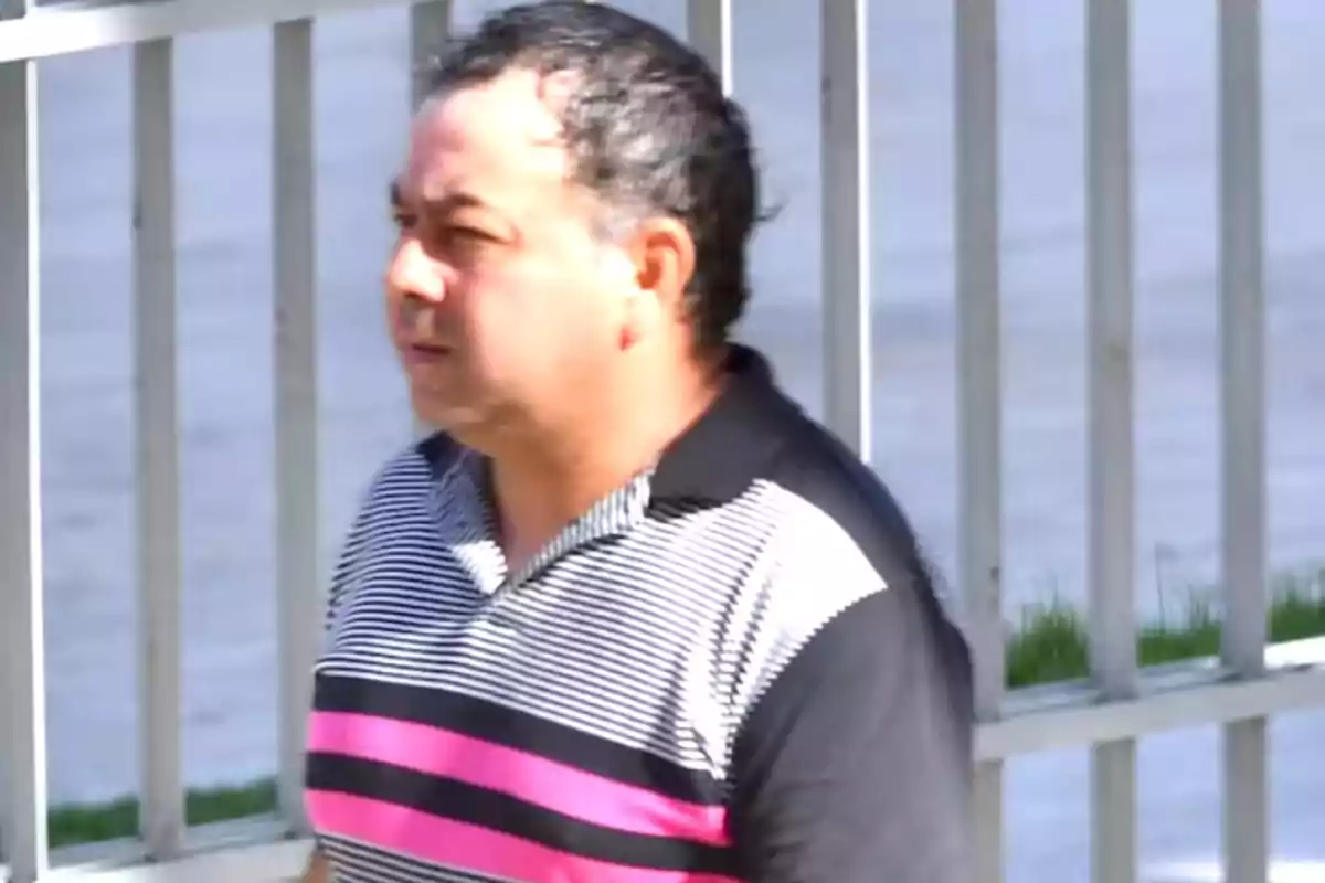 Man with short dark hair wearing a white, black, and pink striped t-jersey walking outdoors next to a metal fence. Man with short dark hair wearing a white, black, and pink striped t-jersey walking outdoors next to a metal fence.
