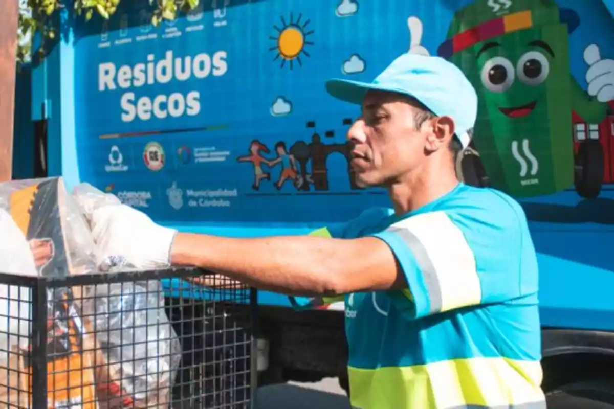 Trabajador de recolección de residuos secos con uniforme azul y guantes colocando materiales reciclables en un contenedor frente a un camión de reciclaje decorado con dibujos y mensajes sobre el cuidado ambiental