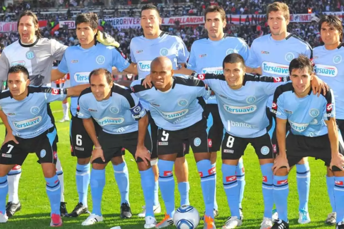Eleven players in light blue and black uniforms pose on the field before a match, with a packed stadium in the background.