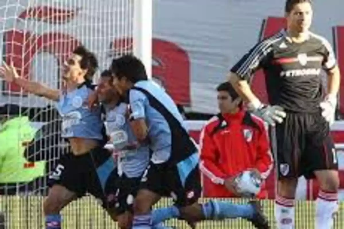Jugadores de un equipo de fútbol con camiseta celeste celebran un gol mientras un arquero con uniforme negro observa la escena