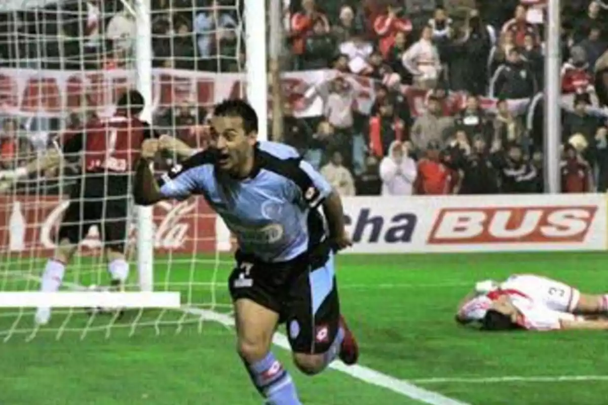 Futbolista con uniforme celeste celebrando un gol mientras un jugador rival está en el suelo y el público observa desde las gradas