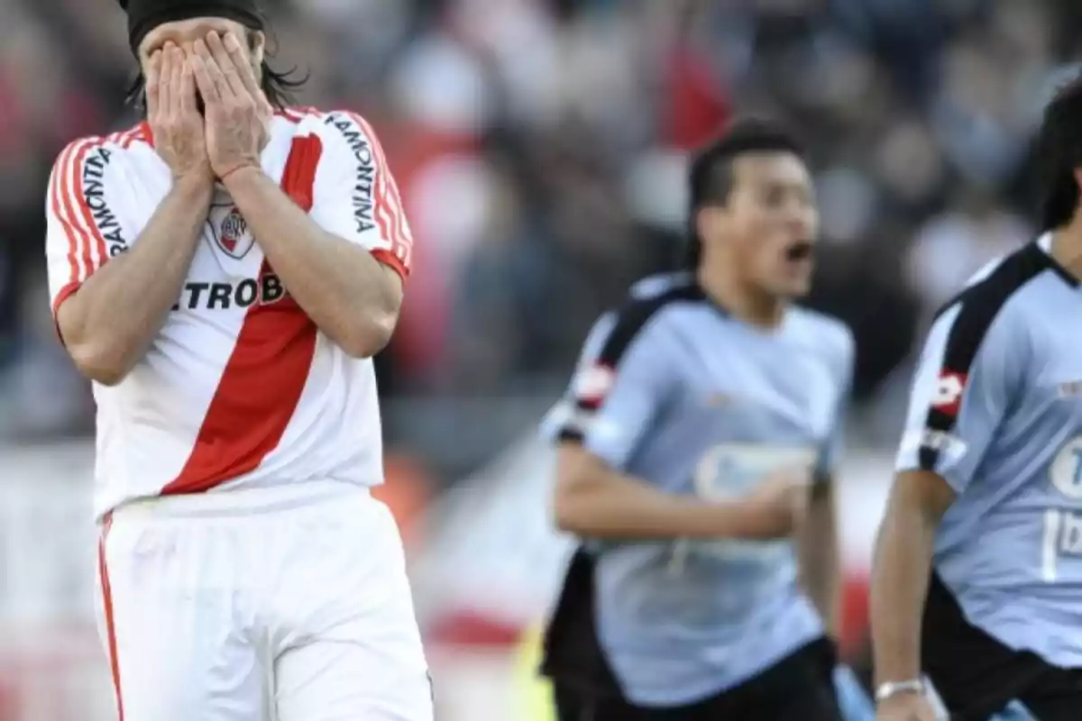 Futbolista con uniforme blanco y franja roja cubriéndose el rostro con las manos mientras jugadores rivales celebran al fondo