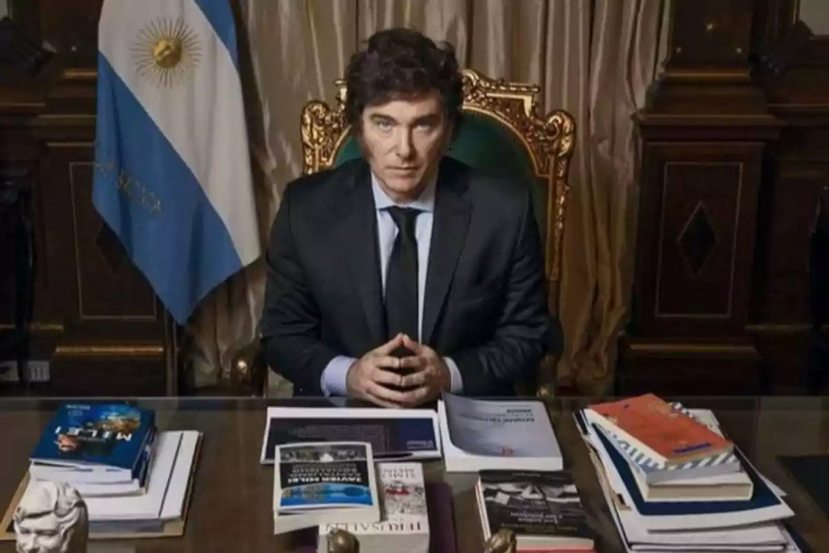 Hombre de traje sentado en un escritorio con varios libros y documentos, al fondo una bandera de Argentina y una silla de respaldo dorado