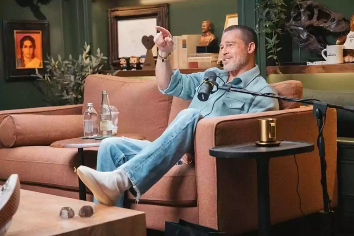Hombre sentado en un sofá color marrón participando en una entrevista con micrófono, rodeado de botellas, una taza y decoración en una sala acogedora Hombre sentado en un sofá color marrón participando en una entrevista con micrófono, rodeado de botellas, una taza y decoración en una sala acogedora