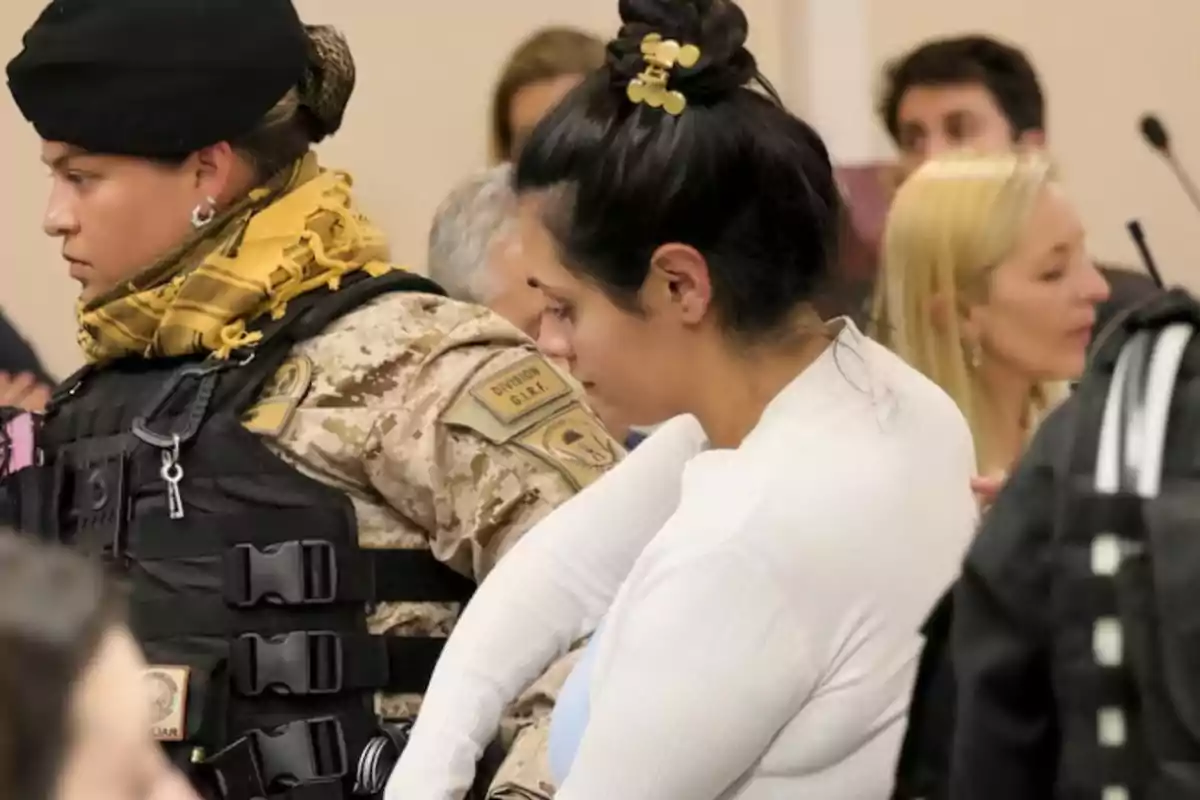 Una mujer escoltada por una agente de seguridad vestida con uniforme táctico en una sala con varias personas al fondo