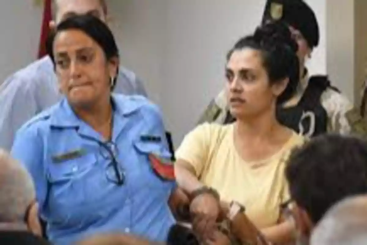 Una mujer con camiseta amarilla esposada es escoltada por una oficial de policía en uniforme azul mientras varias personas observan Una mujer con camiseta amarilla esposada es escoltada por una oficial de policía en uniforme azul mientras varias personas observan
