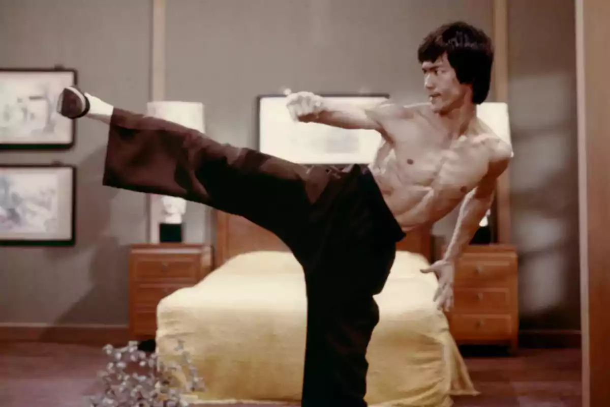 Shirtless man performing a high kick in a room with a bed and wooden furniture Shirtless man performing a high kick in a room with a bed and wooden furniture