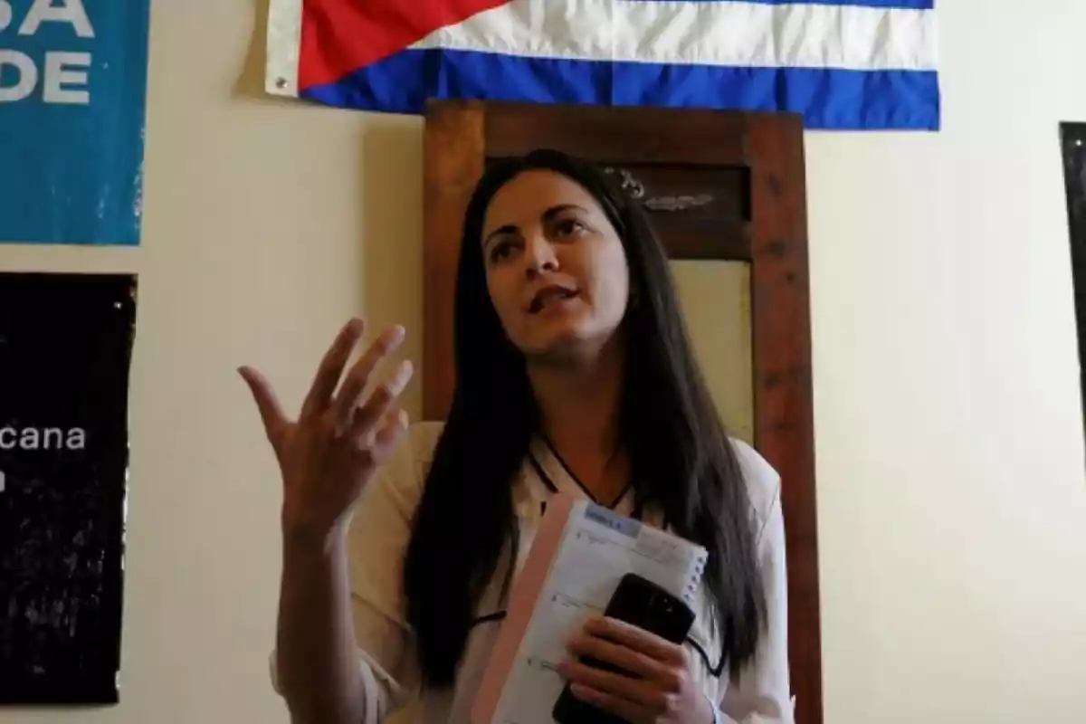 Mujer de cabello largo y oscuro hablando mientras sostiene una libreta y un teléfono móvil, con una bandera de fondo en la pared.