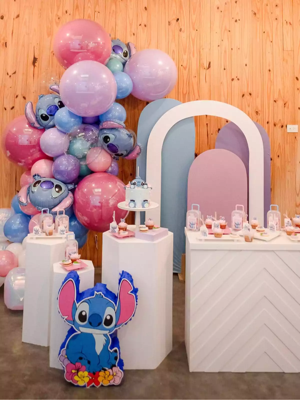 Stitch-themed kids’ party decoration featuring pastel-colored balloons, Stitch figures, tables with cupcakes and sweets, and a wooden backdrop with decorative arches in blue and pink. Stitch-themed kids’ party decoration featuring pastel-colored balloons, Stitch figures, tables with cupcakes and sweets, and a wooden backdrop with decorative arches in blue and pink.